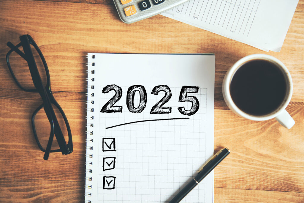 Medical Device Resolutions This image features a notepad with "2025" written boldly at the top, accompanied by a checklist containing three checked boxes. Nearby, there is a pair of eyeglasses, a cup of coffee, a pen, and a calculator, all arranged on a wooden table.