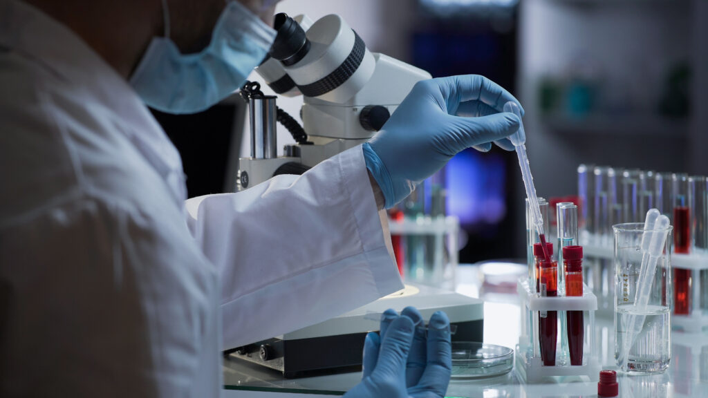 A scientist wearing a white lab coat, blue gloves, and a face mask is using a pipette to transfer liquid into a test tube filled with red fluid. The laboratory setting includes a microscope, multiple test tubes in a rack, and a beaker with pipettes in the background. The environment is well-lit, suggesting a clinical or research laboratory.