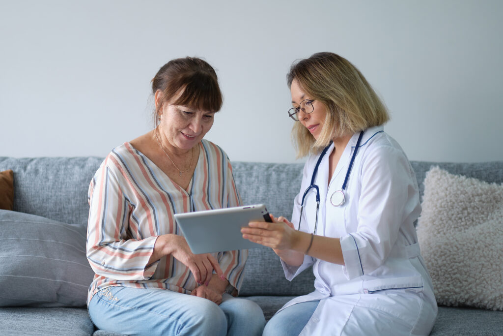 Female professional doctor showing medical test result explaining prescription using digital tablet app visiting senior woman patient at home sitting on sofa. Elderly people healthcare tech concept