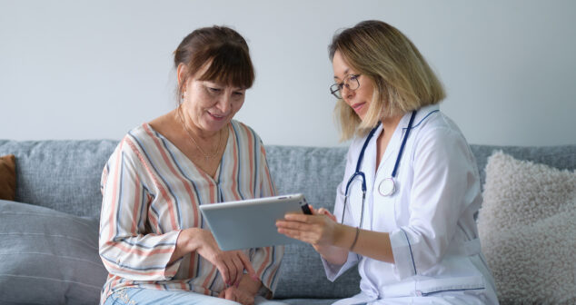 Female professional doctor showing medical test result explaining prescription using digital tablet app visiting senior woman patient at home sitting on sofa. Elderly people healthcare tech concept