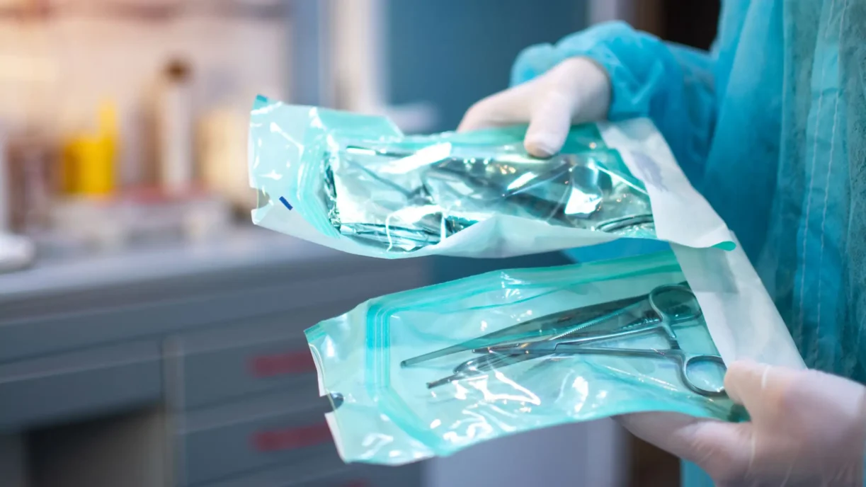 Gloved technician handling sterile packaged medical instruments prepared for radiation sterilization validation.