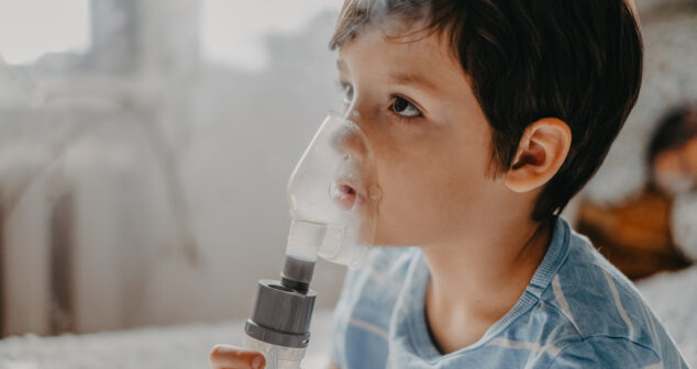 A young child wearing a striped blue shirt uses a nebulizer mask for respiratory treatment. The transparent mask covers their nose and mouth, with visible vapor coming from the device. The child looks upwards while holding the nebulizer, and the background is softly blurred, suggesting a home setting.