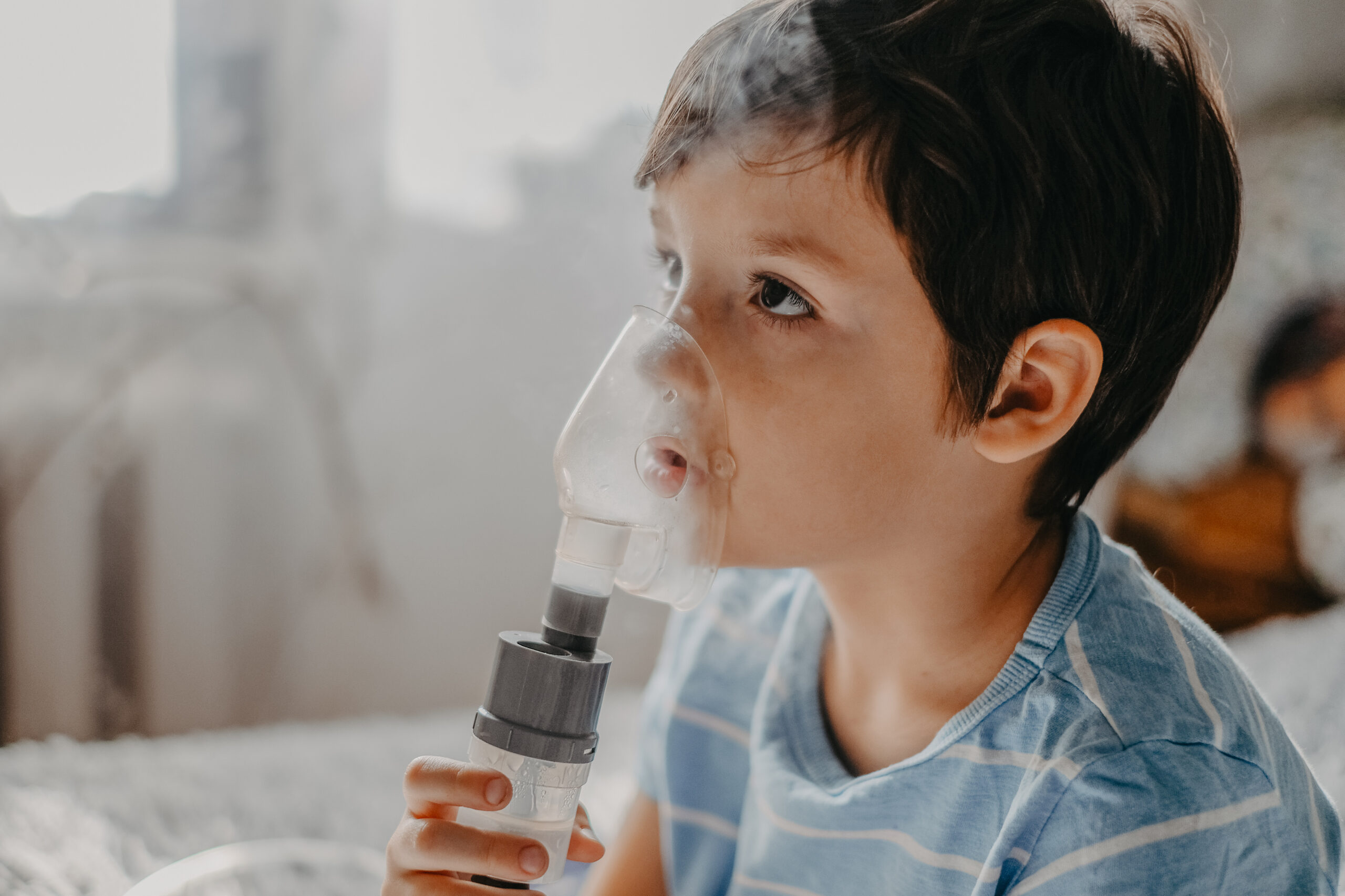 A young child wearing a striped blue shirt uses a nebulizer mask for respiratory treatment. The transparent mask covers their nose and mouth, with visible vapor coming from the device. The child looks upwards while holding the nebulizer, and the background is softly blurred, suggesting a home setting.