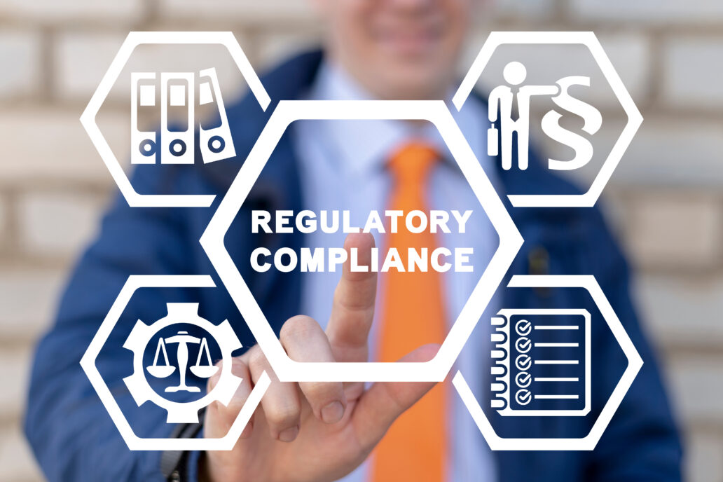 A businessman wearing a blue suit and an orange tie is pointing at a transparent virtual interface with hexagonal icons. The central hexagon displays the words "REGULATORY COMPLIANCE." Surrounding it are four smaller hexagons with white icons, including a scale inside a gear (symbolizing legal and ethical standards), a stack of binders (representing documentation), a checklist (indicating compliance requirements), and a person next to a legal symbol (denoting regulatory oversight). The background is blurred with a brick wall visible.