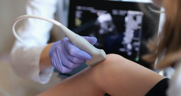 A healthcare professional wearing purple gloves performs an ultrasound examination on a patient's knee using a handheld ultrasound probe. A medical imaging screen displaying the ultrasound results is visible in the background.