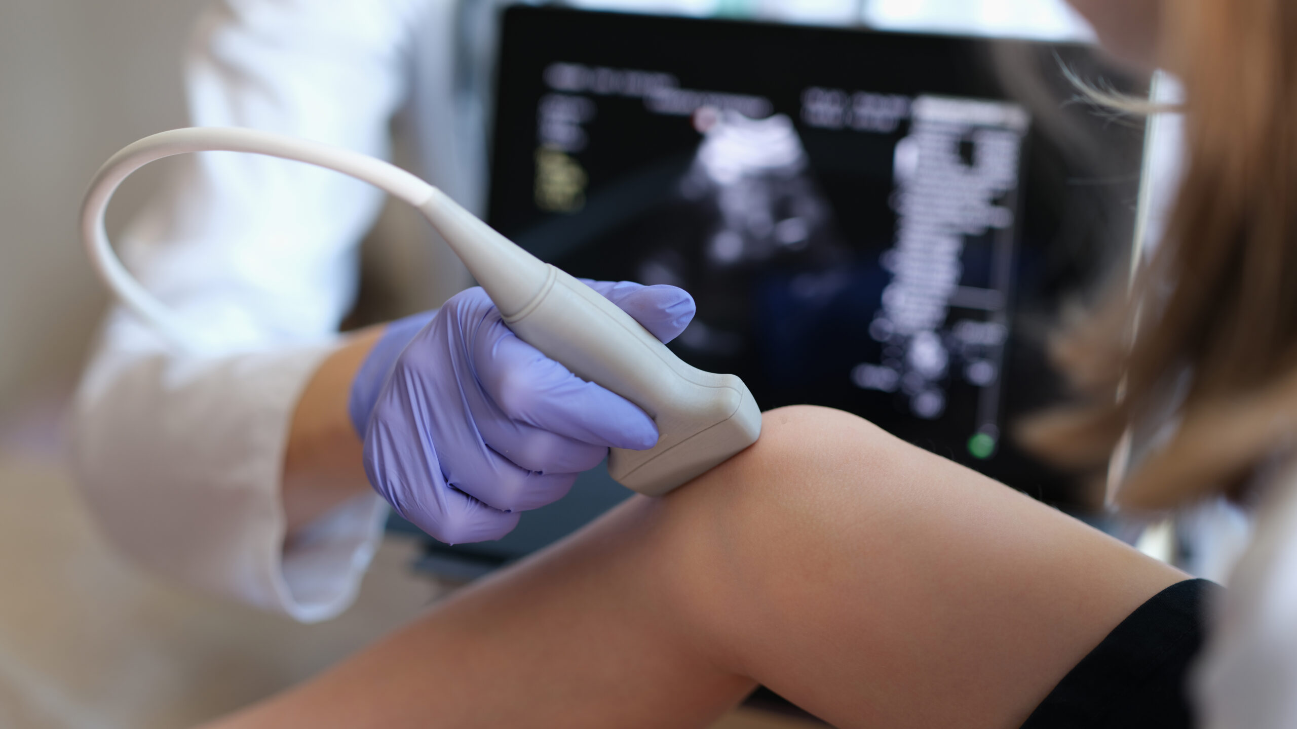 A healthcare professional wearing purple gloves performs an ultrasound examination on a patient's knee using a handheld ultrasound probe. A medical imaging screen displaying the ultrasound results is visible in the background.