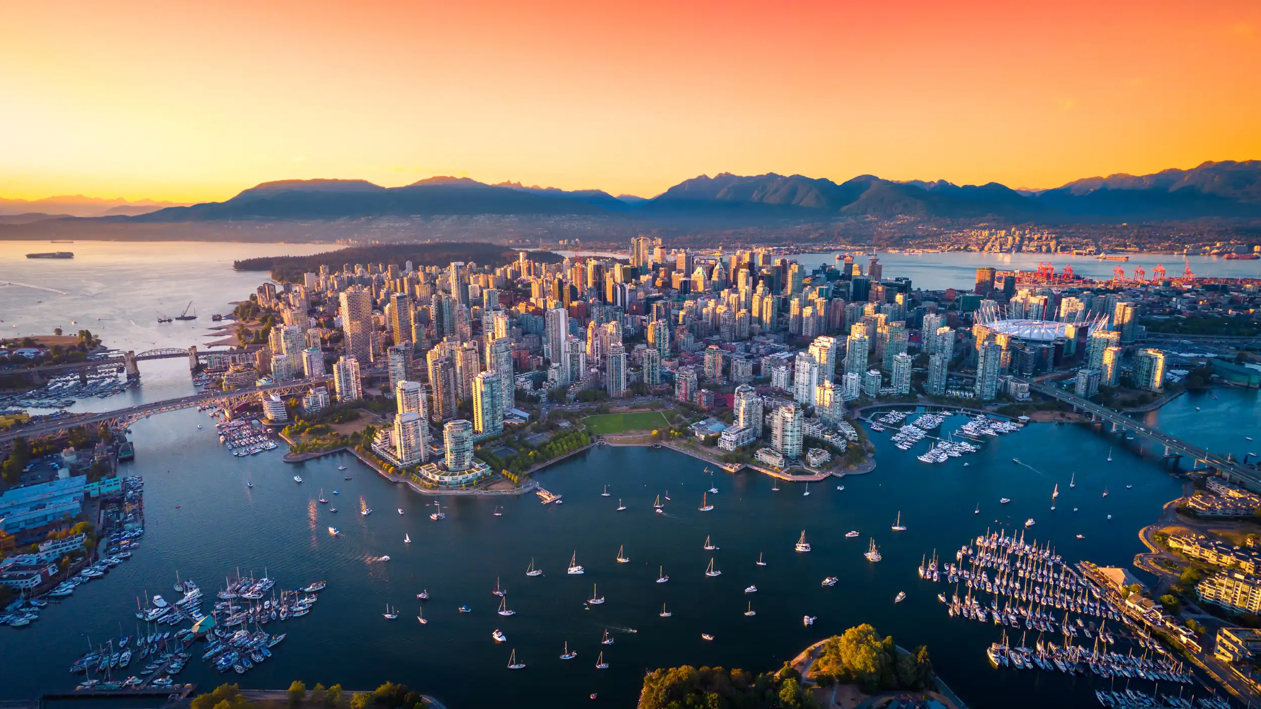 Aerial view of downtown Vancouver at sunset with waterfront and mountains in the background.