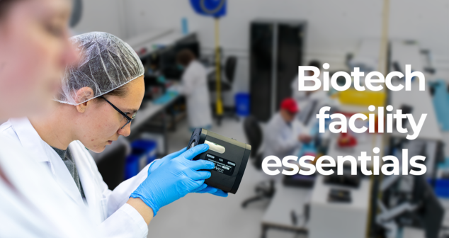 A close-up of a scientist in a white lab coat, wearing a hairnet and blue gloves, examining a device in a biotech laboratory. In the blurred background, several other scientists in lab coats are working at lab benches. The text overlay reads "Biotech facility essentials.