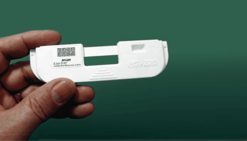 Hand holding a white handheld IVD cartridge labeled with a QR code and testing information against a green background.