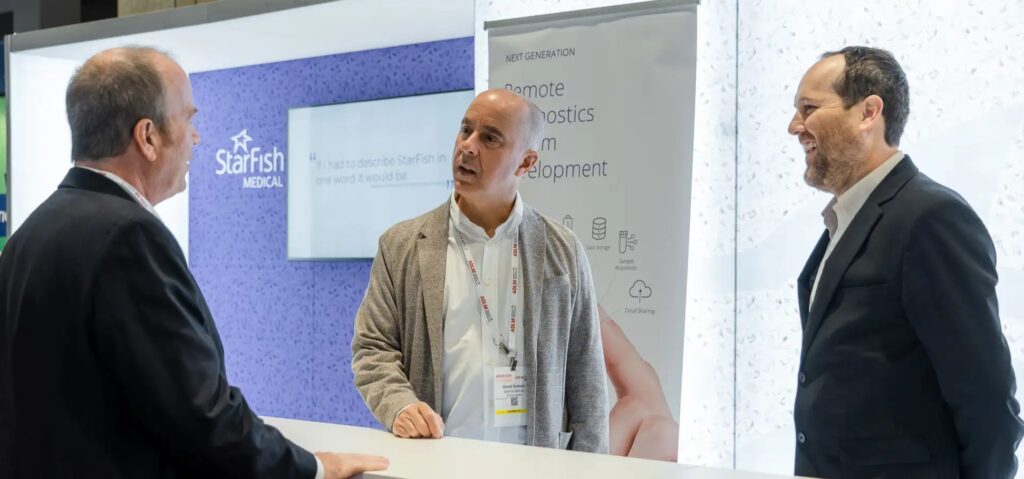 Three professionals talk at a StarFish Medical trade show booth, with signage highlighting remote diagnostics and medical device development in the background.