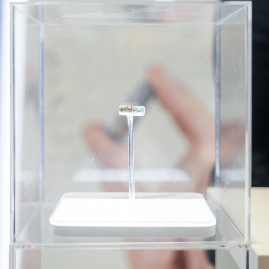 The image shows a small, cylindrical device encased in a clear capsule, displayed on a transparent stand within a glass enclosure. The background is blurred, highlighting the miniature device as the focal point, with a modern, well-lit presentation.