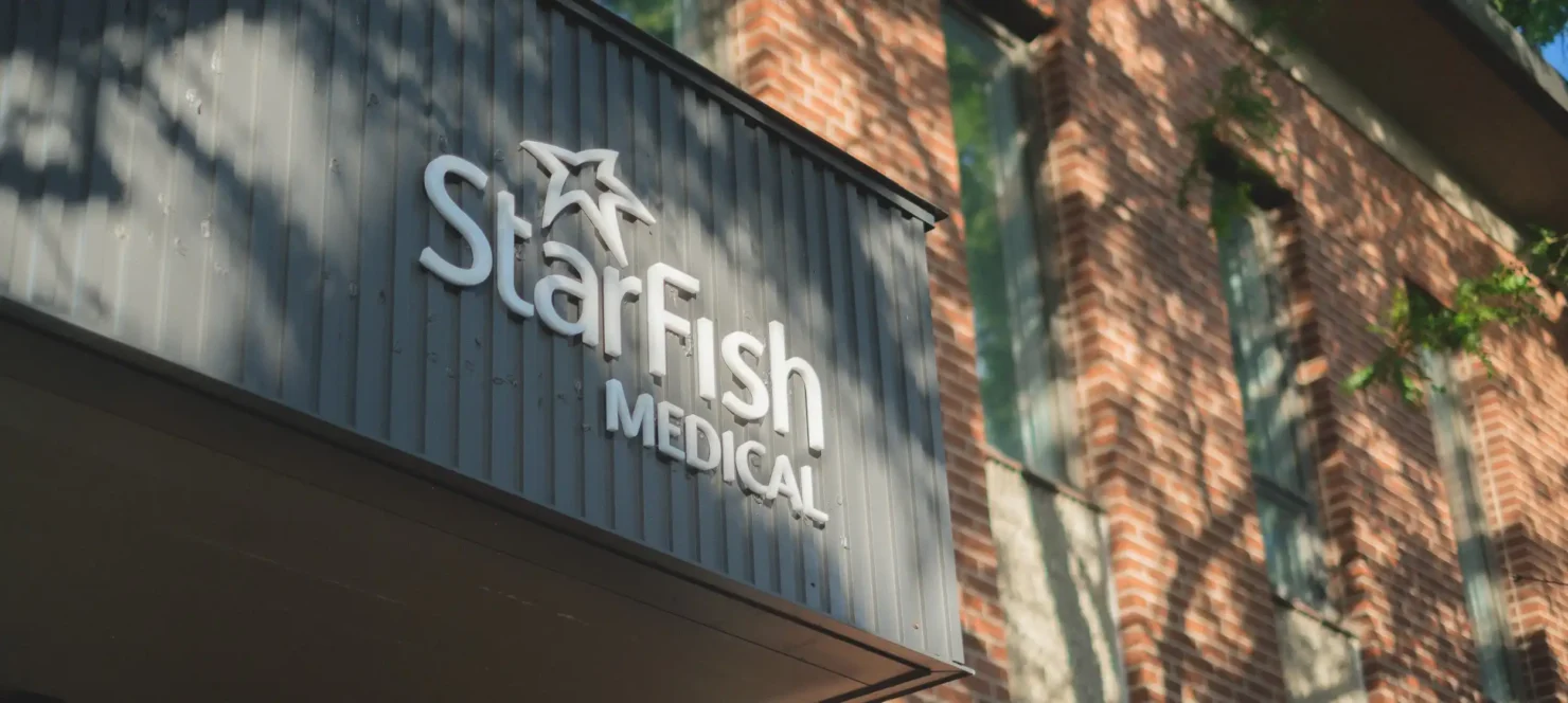 Exterior view of StarFish Medical Toronto office sign on a gray building facade with a brick wall and window in the background.
