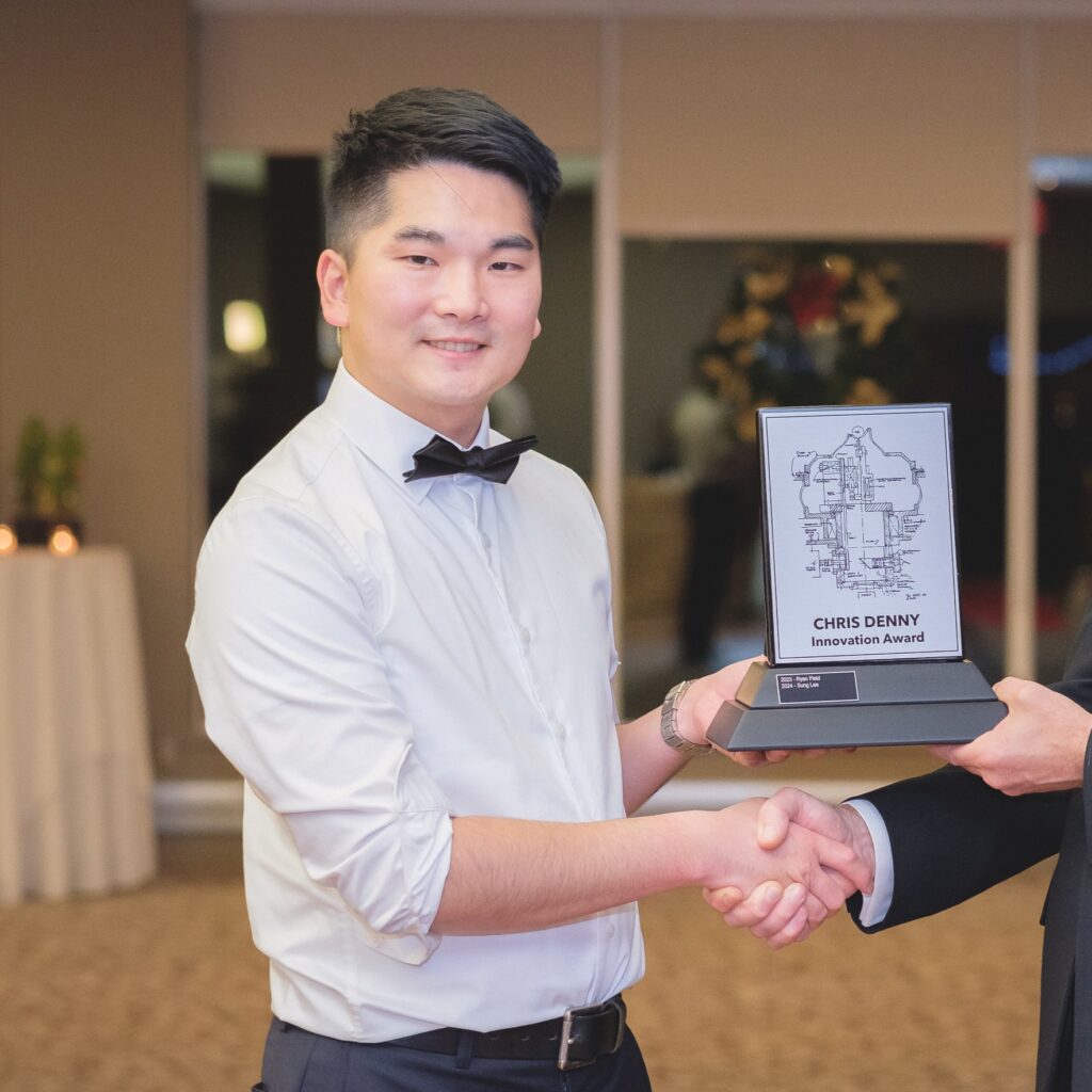 A person wearing a formal white shirt with rolled-up sleeves and a black bow tie. They are smiling while holding an award titled "Chris Denny Innovation Award." The award has a design resembling a technical or mechanical diagram. The person is shaking hands with someone whose arm is partially visible in the frame, wearing a black suit. The background suggests a formal event setting with dim lighting and some decorations.