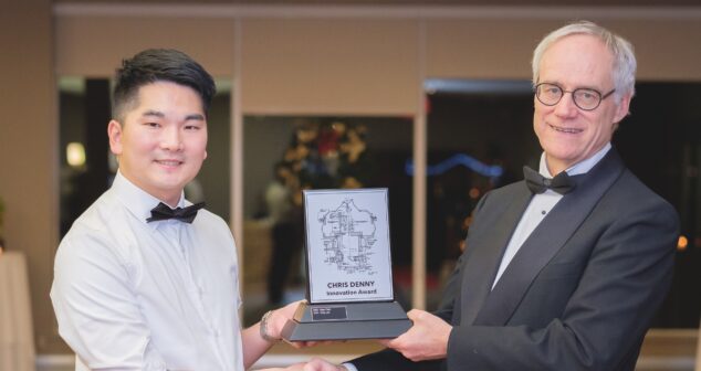 Two individuals dressed formally, with one presenting an award that says "Chris Denny Innovation Award." The recipient is smiling and wearing a bow tie.