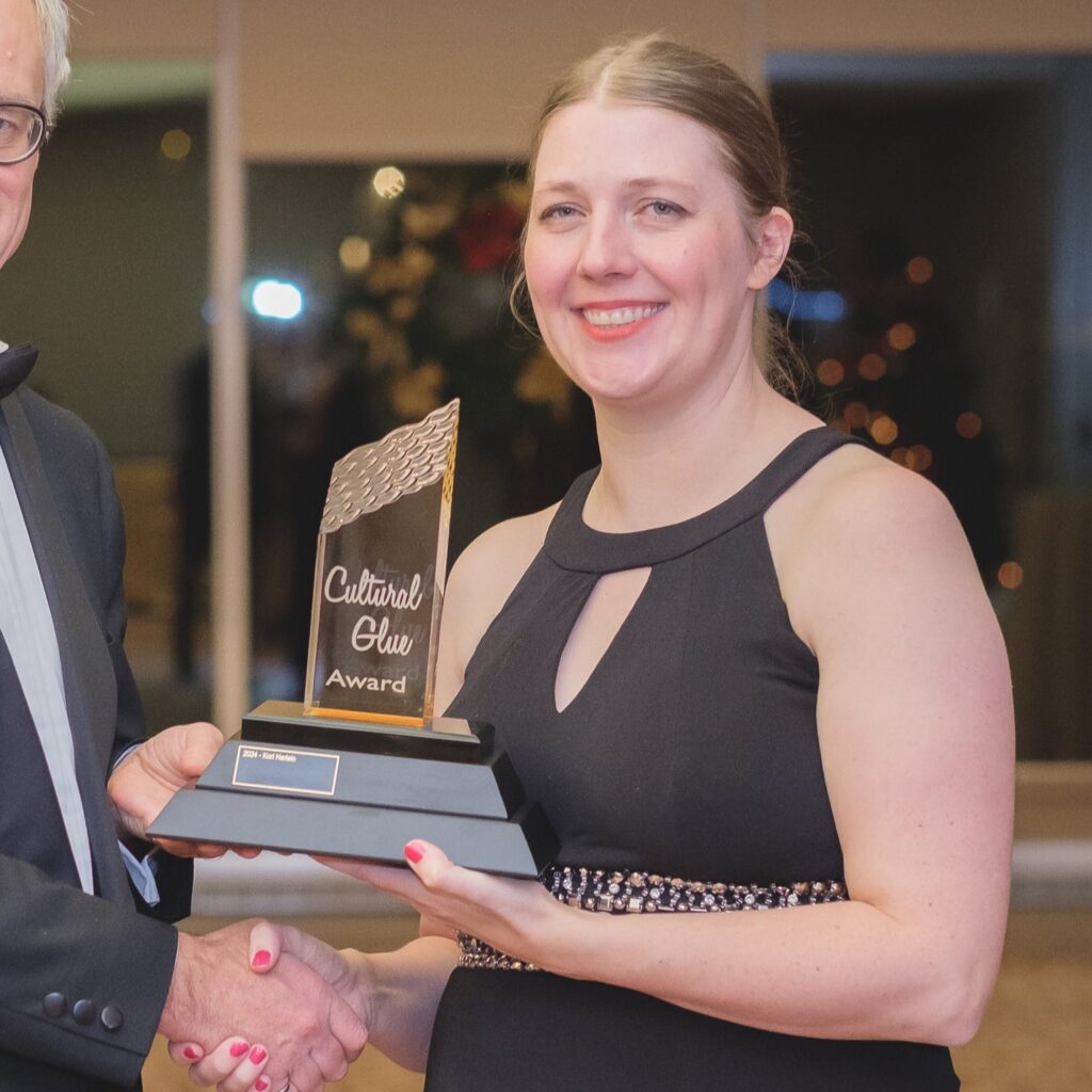 A woman in a black formal dress holding a trophy labeled "Cultural Glue Award" and smiling.