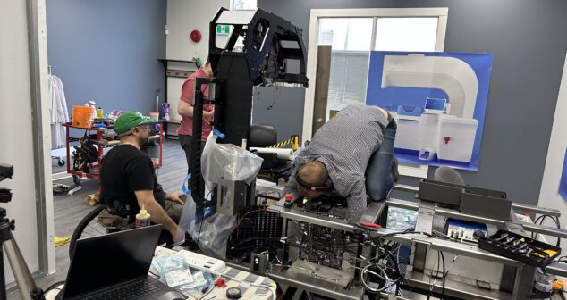 Three engineers are assembling or troubleshooting a large industrial or medical device prototype in a workshop. One person is seated in a wheelchair, another is standing behind the structure, and a third is kneeling on top of the machine for internal access. The workspace includes tools, laptops, electronic parts, and a large diagram of the device on the wall.