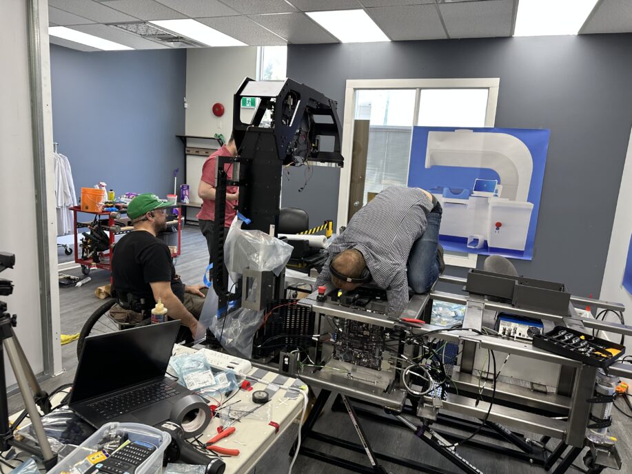 Three engineers are assembling or troubleshooting a large industrial or medical device prototype in a workshop. One person is seated in a wheelchair, another is standing behind the structure, and a third is kneeling on top of the machine for internal access. The workspace includes tools, laptops, electronic parts, and a large diagram of the device on the wall.
