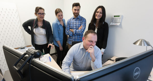 device product development Accessibility: man pondering a double screen computer with four people looking from behind him.