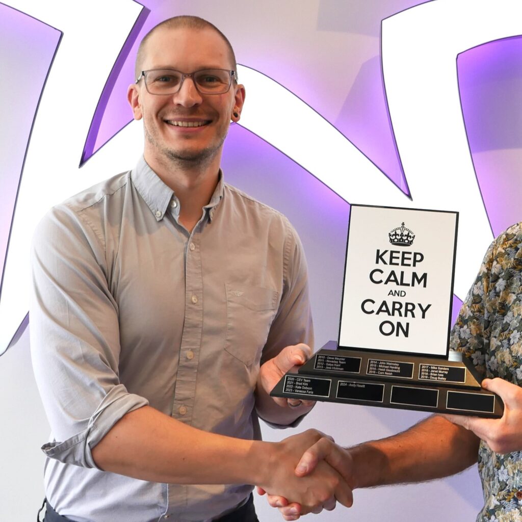 A man wearing glasses and a light shirt holding a plaque labeled "Keep Calm and Carry On" while shaking hands with another individual.