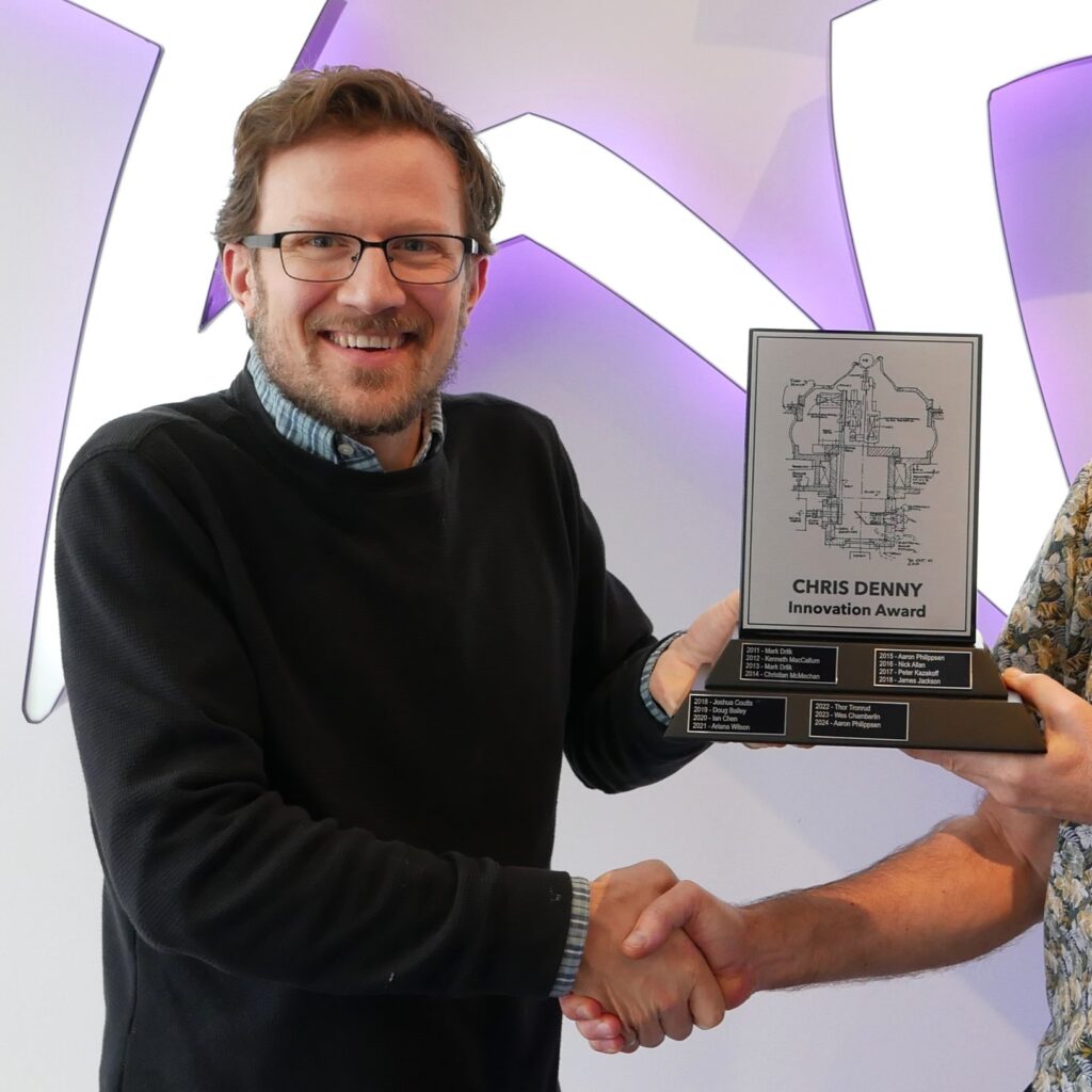 A man smiling and holding an award with "Chris Denny Innovation Award" written on it, shaking hands with another person.