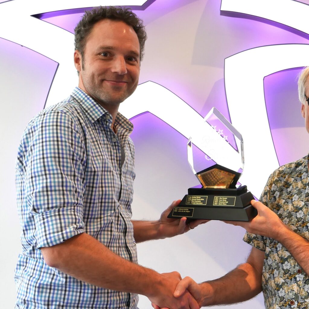 A man in a checkered shirt shaking hands while holding an award with a star-shaped backdrop in the background.