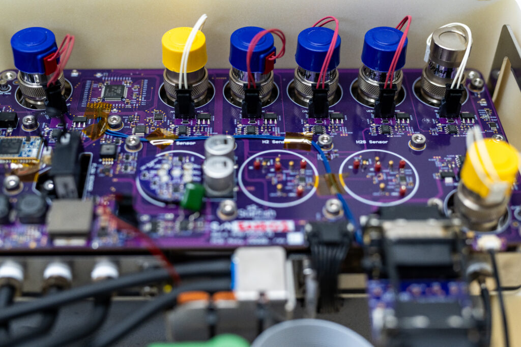 Close-up of a printed circuit board (PCB) with multiple connectors, sensors, and electronic components. The board has a purple solder mask with various capacitors, resistors, and ICs mounted. Colored caps on the connectors indicate different sensor inputs, with red and white wires attached. Visible traces and test points suggest a complex design, likely for a medical or industrial application.