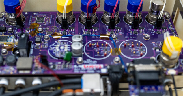 Close-up of a printed circuit board (PCB) with multiple connectors, sensors, and electronic components. The board has a purple solder mask with various capacitors, resistors, and ICs mounted. Colored caps on the connectors indicate different sensor inputs, with red and white wires attached. Visible traces and test points suggest a complex design, likely for a medical or industrial application.