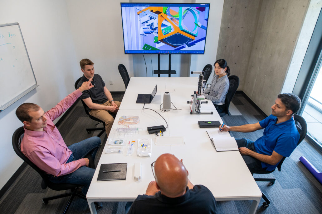 underlying problem Accessibility: engineers seated at table disucssing image on large monitor mounted on wall at the far end of the table.