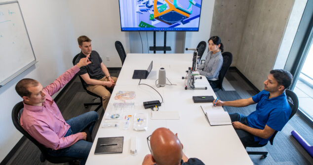 underlying problem Accessibility: engineers seated at table disucssing image on large monitor mounted on wall at the far end of the table.