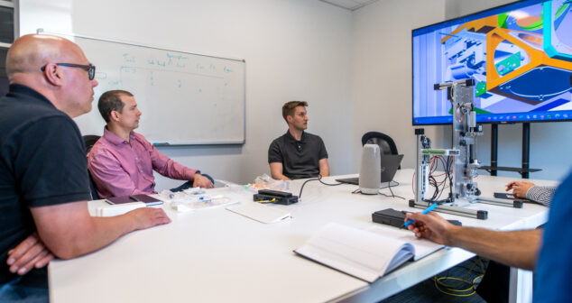 ‘Fee for Service’ Model Team of people gathered around conference table discussing image CAD design on monitor at back wall of room