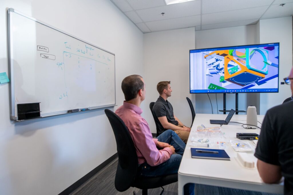 A team of professionals sitting in a conference room, engaged in a discussion. A large screen displays a colorful 3D design, while a whiteboard on the wall shows notes and diagrams. The table is equipped with laptops, notepads, and engineering tools, emphasizing a collaborative work environment.