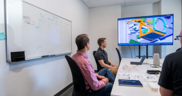 A team of professionals sitting in a conference room, engaged in a discussion. A large screen displays a colorful 3D design, while a whiteboard on the wall shows notes and diagrams. The table is equipped with laptops, notepads, and engineering tools, emphasizing a collaborative work environment.