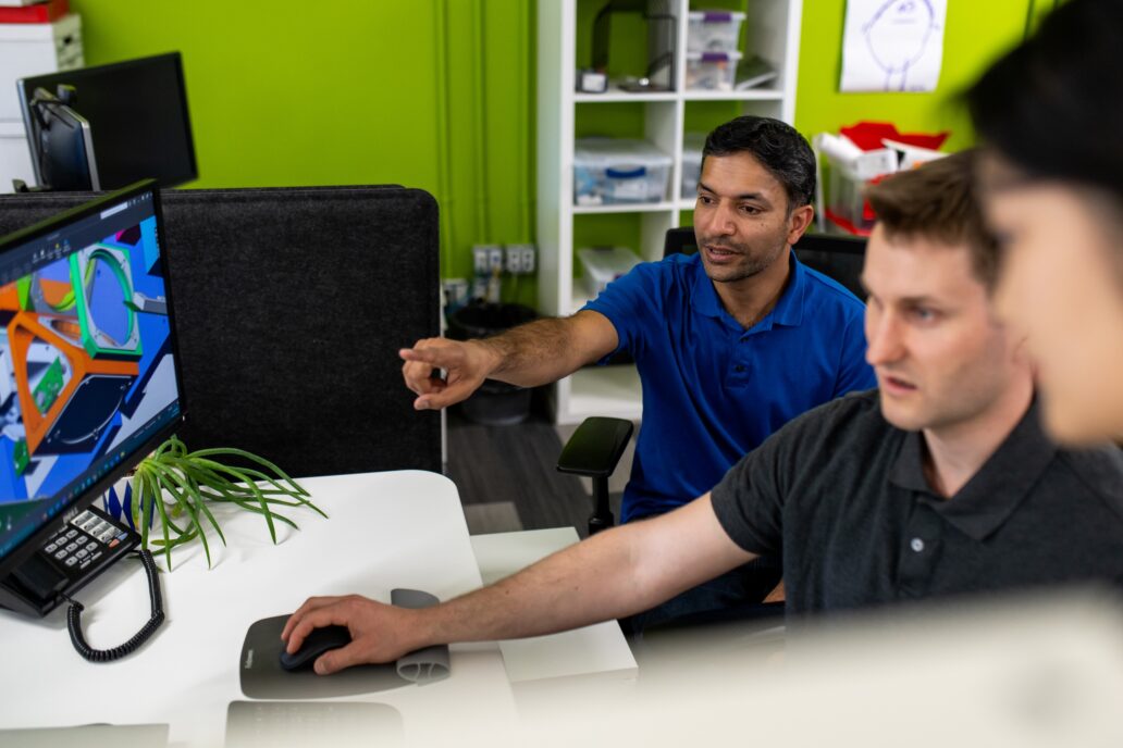 Three team members collaborating at a workstation in a modern office environment. One person in a blue shirt is pointing at a computer screen displaying a colorful 3D model, while another operates the mouse. The workspace features a green wall, shelving with supplies, and a small plant on the desk, creating a dynamic and engaging atmosphere.