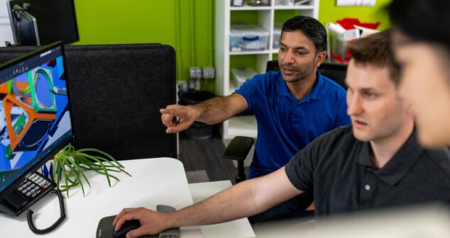 Three team members collaborating at a workstation in a modern office environment. One person in a blue shirt is pointing at a computer screen displaying a colorful 3D model, while another operates the mouse. The workspace features a green wall, shelving with supplies, and a small plant on the desk, creating a dynamic and engaging atmosphere.