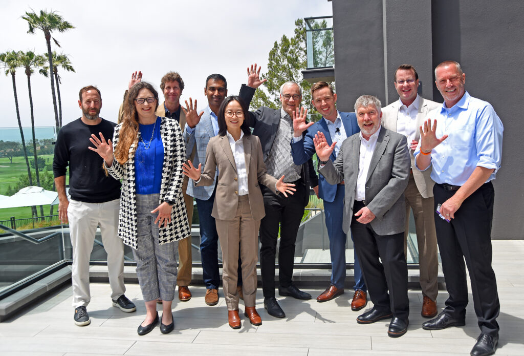 A group of speakers at Medical Device Playbook Newport Beach 2025 pose outdoors on a terrace with palm trees and a golf course in the background. Most are waving with one hand raised. They are dressed in business casual and formal attire, suggesting a networking or conference setting.