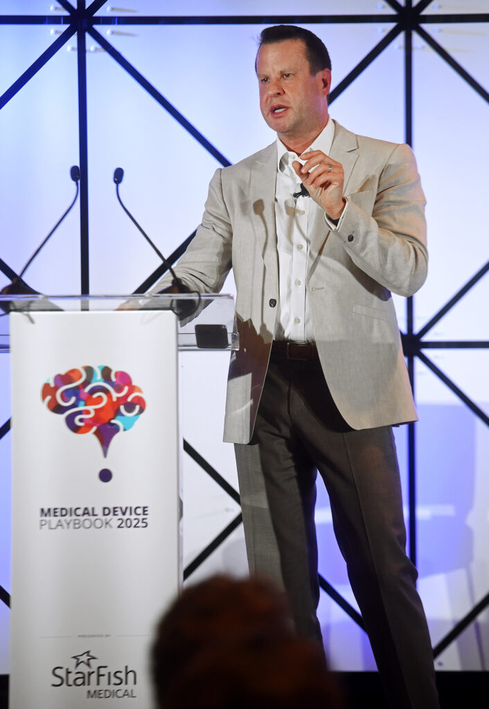 Ken Mariash in a beige blazer speaks at a podium labeled "Medical Device Playbook Newport Beach 2025." The background features a geometric black-and-white pattern. He gestures with his right hand while addressing the audience, conveying a confident and engaging presence.