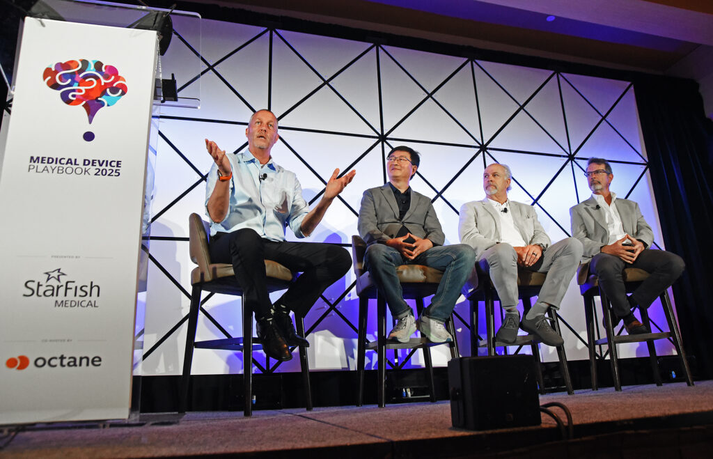 Mike Hill, Henry Yan, John Guidon, and Len Brandt sit on a stage under the "Medical Device Playbook 2025" banner. The man on the far left gestures animatedly while speaking, and the others listen attentively. The setup includes high chairs and a geometric backdrop with blue and purple lighting.