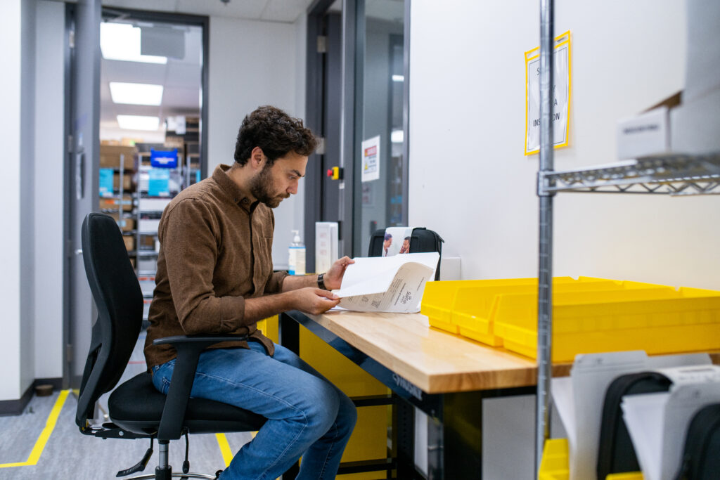 A man seated at a workstation in a lab or manufacturing environment, reading a document. He is surrounded by yellow storage bins, shelves, and labeled areas, with a production or inventory space visible in the background.
