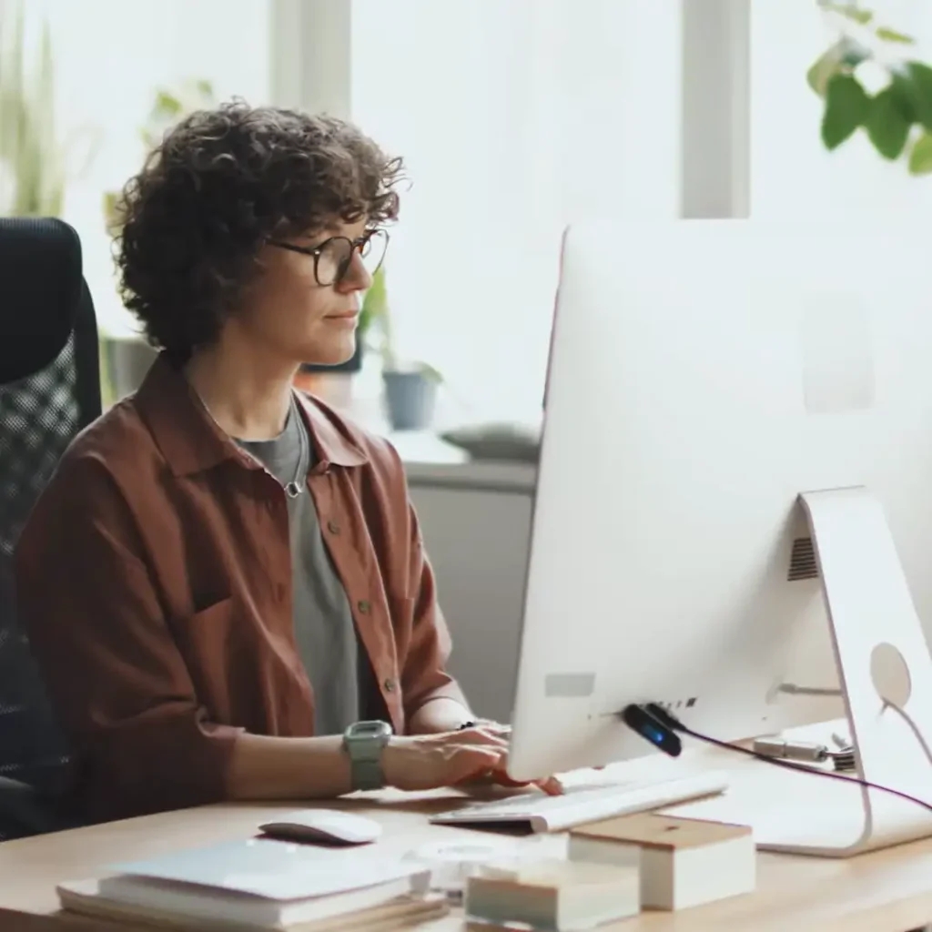 ELLA AI FDA regulatory research tool A person with curly hair and glasses sitting at a desk, typing on a computer in a brightly lit office with plants in the background. The desk has office supplies and a cup of coffee.