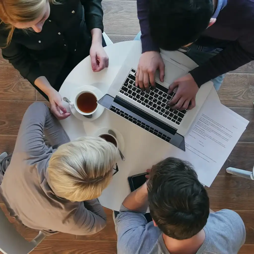 ELLA AI FDA regulatory research tool A group of four people sitting around a circular table with a laptop open, discussing something. Papers, coffee cups, and a smartphone are on the table. The scene conveys collaboration.