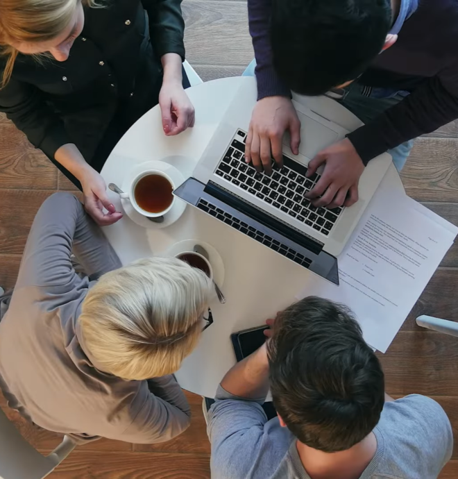 A group of four people sitting around a circular table with a laptop open, discussing something. Papers, coffee cups, and a smartphone are on the table. The scene conveys collaboration.