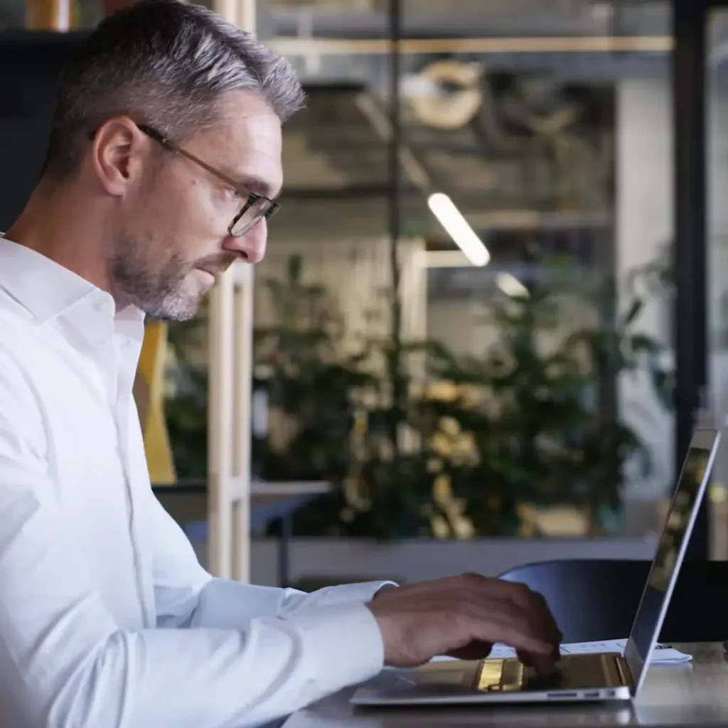 A man with short gray hair and glasses, wearing a white button-up shirt, is working on a laptop at a desk in a modern office with plants and industrial-style decor in the background.