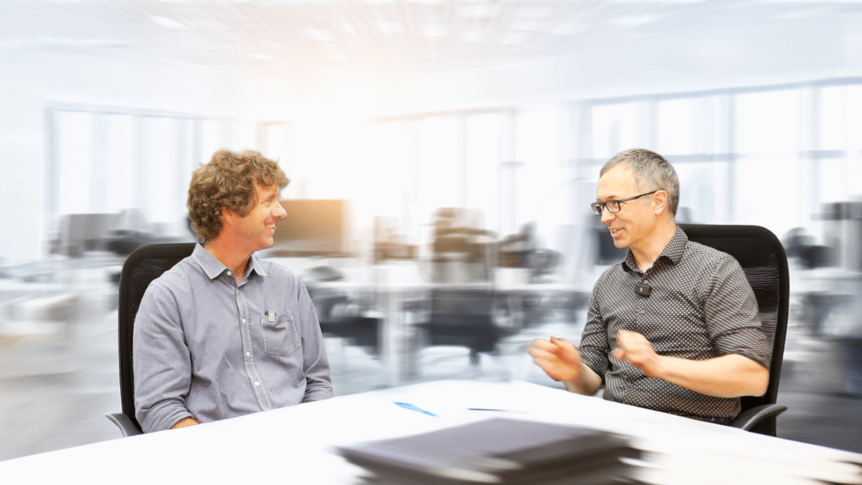 Paul Charlebois and Eric Olson are seated across from each other at a white table in a modern office setting. The man on the left has curly brown hair and is wearing a light blue button-up shirt. He is smiling and looking toward the man on the right, who has short gray hair, glasses, and is wearing a dark patterned shirt with a small microphone clipped to his collar. The background is a bright, blurred open-plan office with desks and chairs, creating a soft focus effect behind the subjects. A blue pen and a blurred stack of folders or documents are on the table in the foreground.