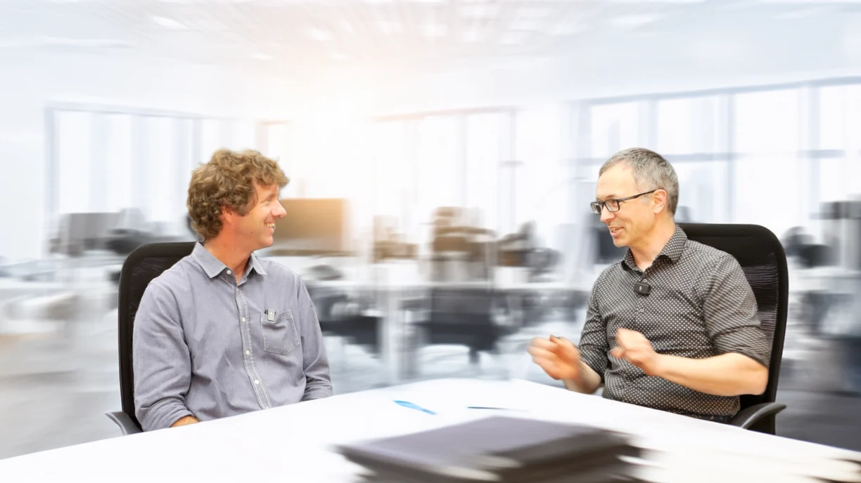 Paul Charlebois and Eric Olson are seated across from each other at a white table in a modern office setting. The man on the left has curly brown hair and is wearing a light blue button-up shirt. He is smiling and looking toward the man on the right, who has short gray hair, glasses, and is wearing a dark patterned shirt with a small microphone clipped to his collar. The background is a bright, blurred open-plan office with desks and chairs, creating a soft focus effect behind the subjects. A blue pen and a blurred stack of folders or documents are on the table in the foreground.