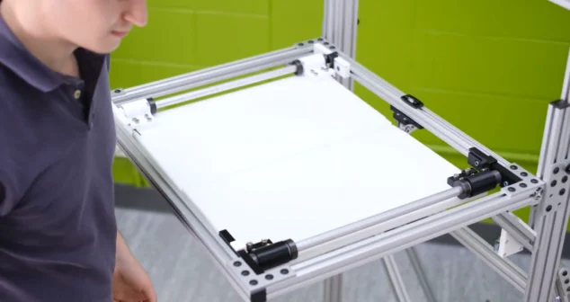 Technician operating a custom aluminum-framed drop test rig with a white platform and green background wall.