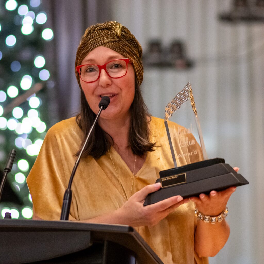 A woman in a gold outfit and red glasses holding a trophy labeled "Cultural Glue Award" while speaking at a podium, with a festive background.