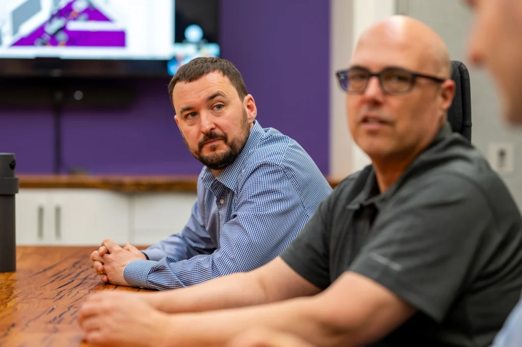 Three men seated around a conference table engaged in a meeting. One man in a blue checkered shirt looks attentively toward a speaker off-camera, while the others listen. A screen with a diagram is visible in the background, and the room features a purple accent wall.