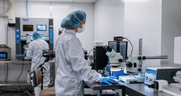 Two laboratory professionals in cleanroom attire, including hair covers, masks, and gloves, working in a high-tech lab. One person is using a microscope at a workstation, while the other operates a machine in the background. The environment is clean and well-organized, featuring advanced scientific equipment and tools.