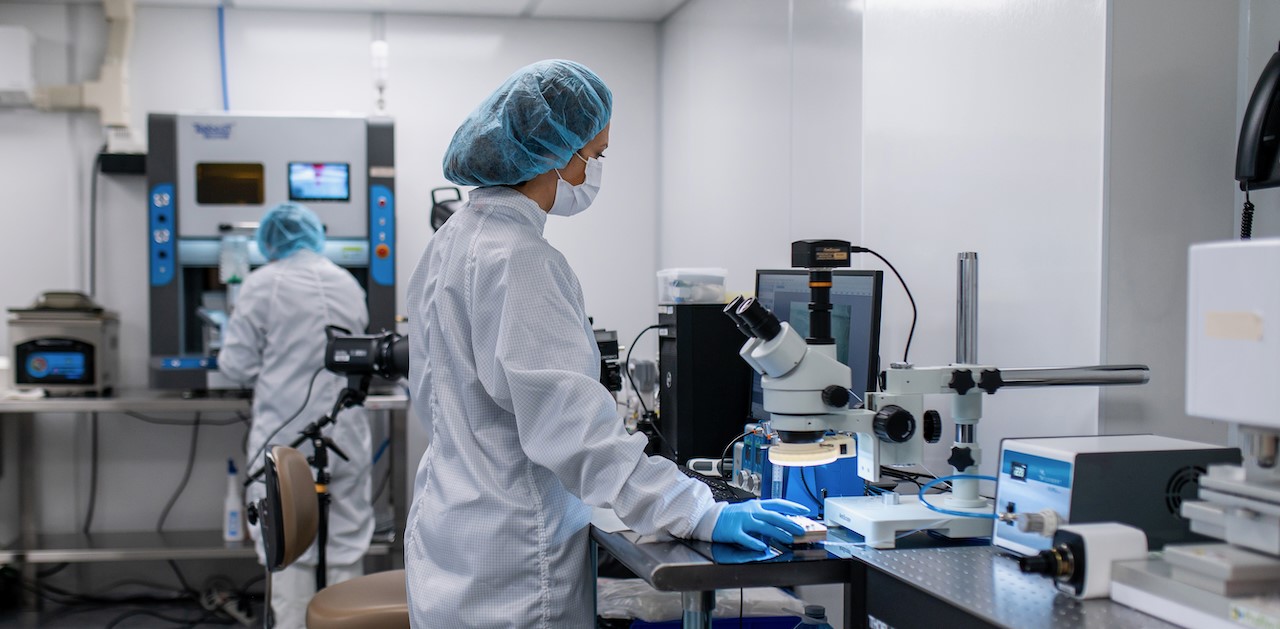 Two laboratory professionals in cleanroom attire, including hair covers, masks, and gloves, working in a high-tech lab. One person is using a microscope at a workstation, while the other operates a machine in the background. The environment is clean and well-organized, featuring advanced scientific equipment and tools.