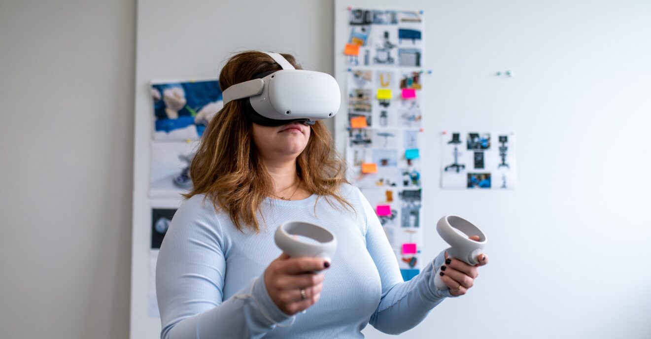 A person wearing a white virtual reality headset and holding two VR controllers stands in a brightly lit room. Behind them, a whiteboard with various images, notes, and colorful sticky notes is visible, suggesting a creative or collaborative workspace.